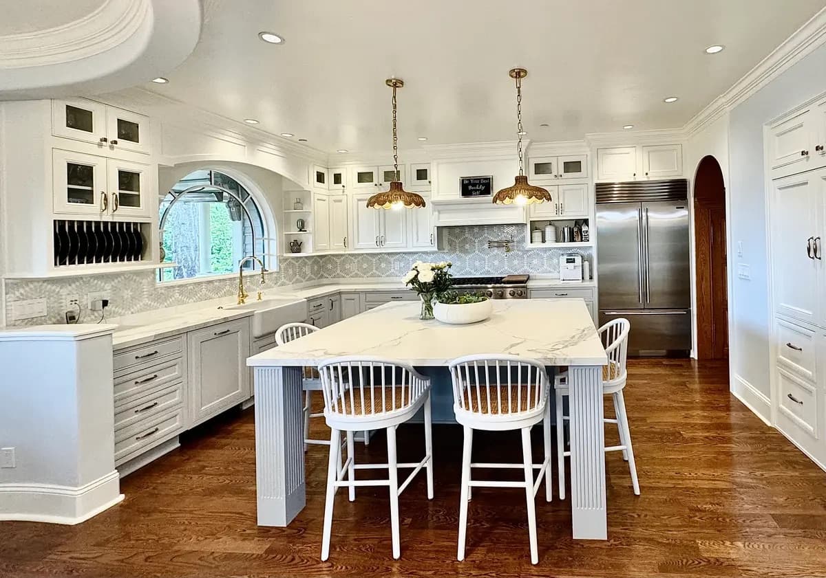 White kitchen with arched window, brass pendants, and a marble island with dining seating