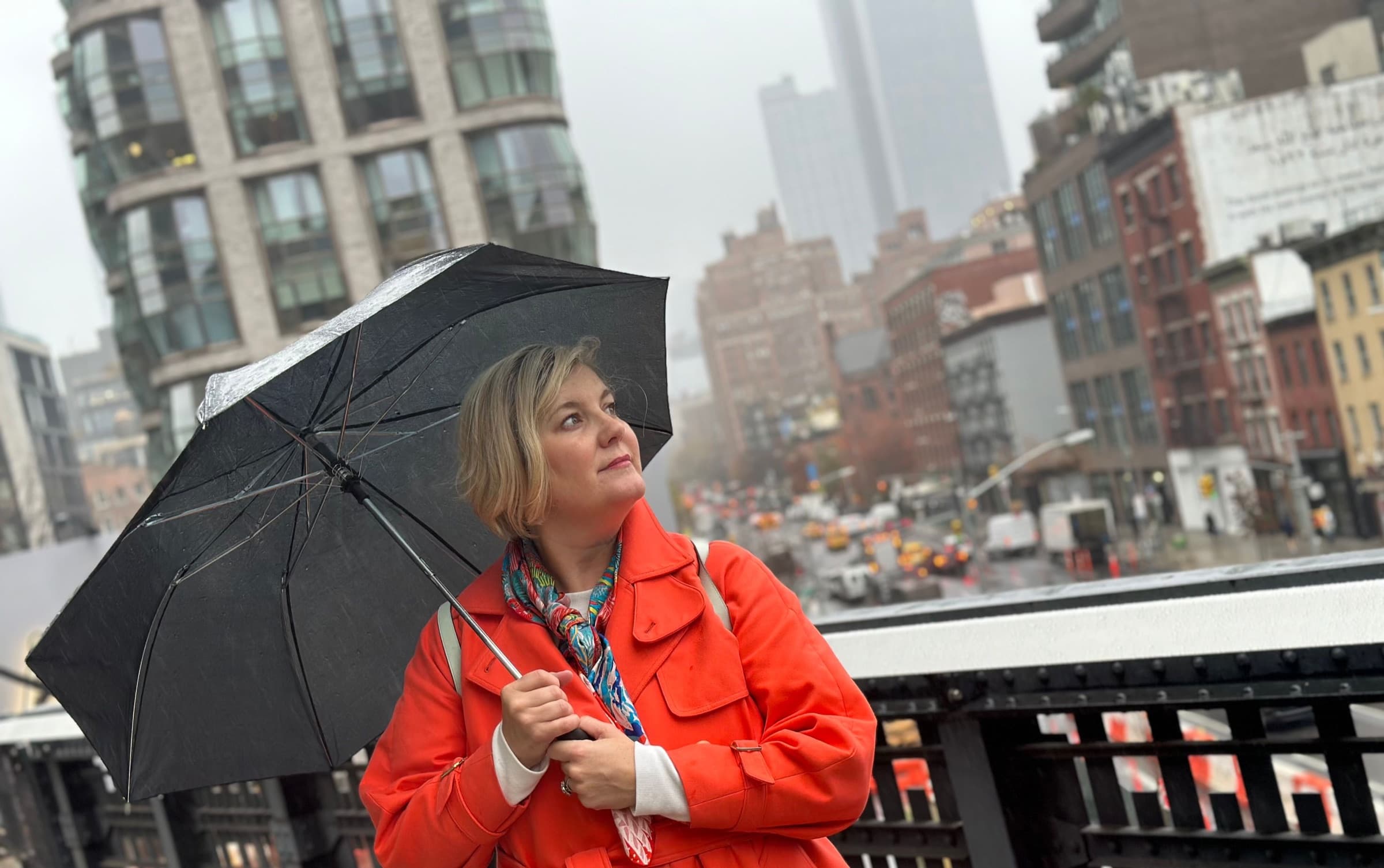 Skye Howell Henley in an orange coat under a black umbrella on a rainy New York street
